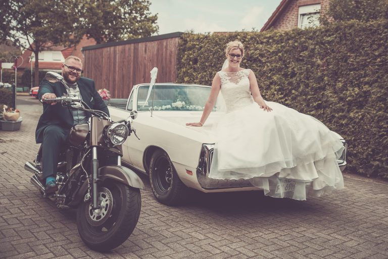 Bräutigam auf einem Motorrad, die Braut sitzt auf dem Kofferraum eines weißen Cabrios