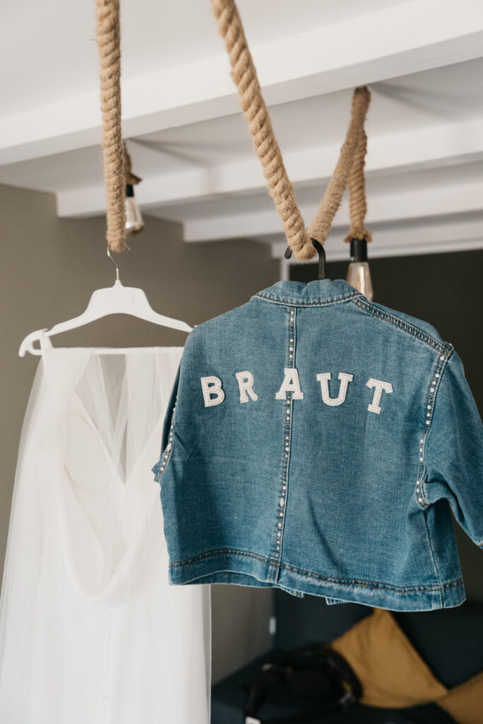 Eine blaue Jeansjacke mit der Aufschrift Braut hängt neben einem weißen Brautkleid an einer Leuchte mit dicken Seilen.