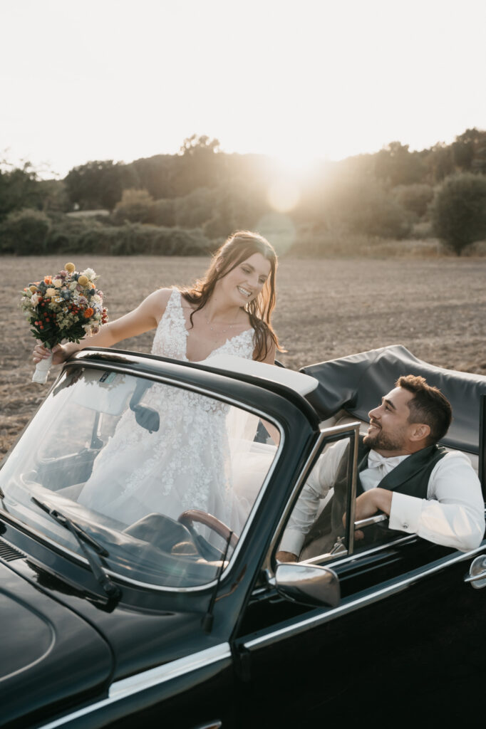Eine Braut steht mit ihrem Blumenstrauß neben einem offenen dunklen Cabrio und lächelt zum Bräutigam auf dem Fahrersitz. Die tief stehende Sonne leuchtet hinter dem Feld.
