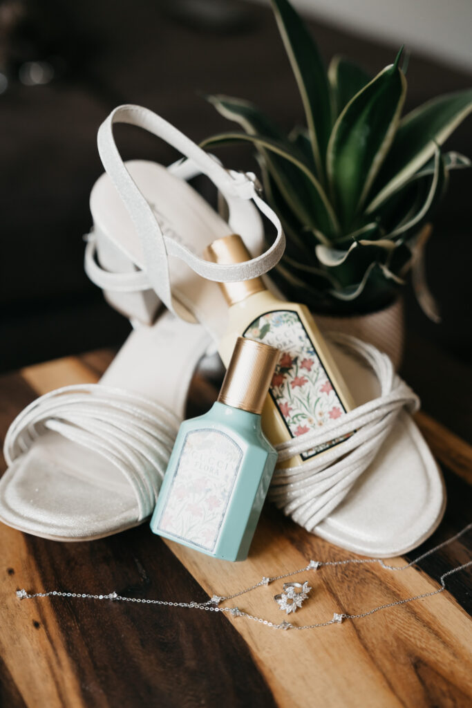 Weiße Riemchensandalen, zwei Parfumflakons und silberfarbener Schmuck liegen auf einem Holztisch. Eine kleine grüne Pflanze steht dahinter.