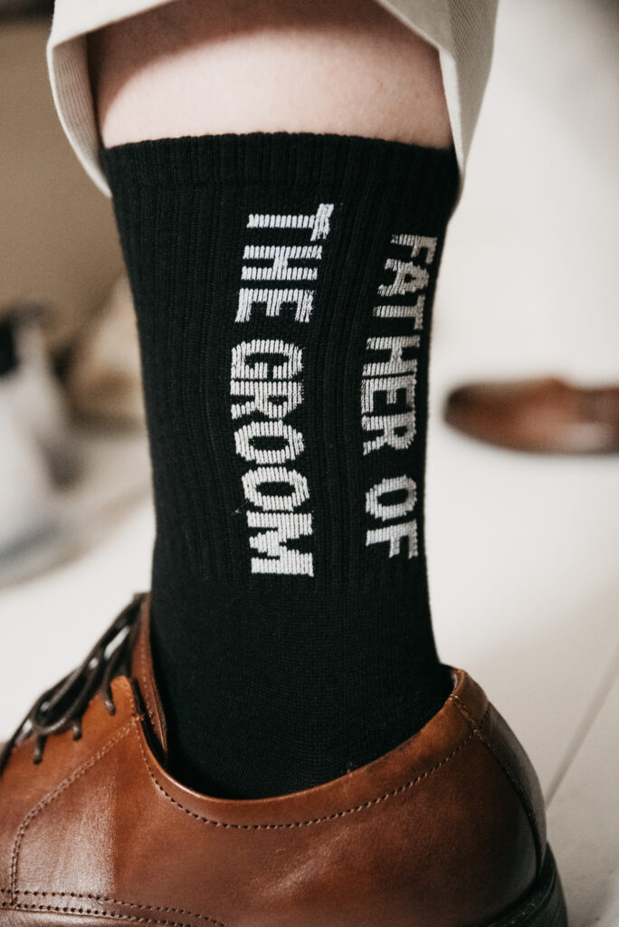 Ein schwarzer Socken trägt die weiße Aufschrift Father of the Groom. Dazu sind ein brauner Lederschuh und der hochgezogene Rand einer hellen Hose zu sehen.