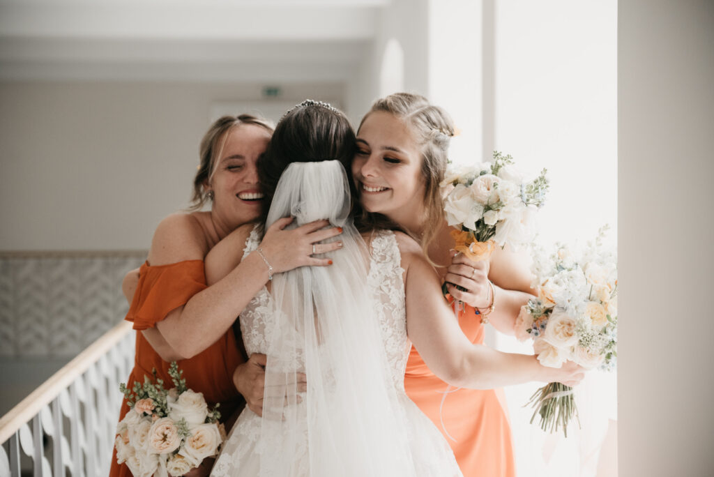 Zwei lächelnde Frauen in orangefarbenen Kleidern umarmen eine Braut mit Schleier. Alle drei halten helle Blumensträuße, weiches Fensterlicht fällt auf sie.