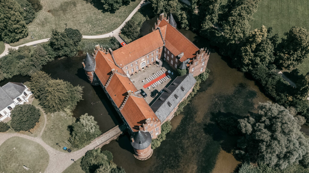 Luftaufnahme eines roten Wasserschlosses mit runden Ecktürmen. Im Innenhof stehen weiße Sitzreihen beidseits eines roten Teppichs, außen liegen Wassergraben und Park.